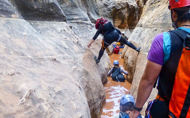 A group of people navigate through a narrow canyon filled with water. Some are wading in the waist-deep water, while another attempts to walk above the water, bracing their feet on either side of the canyon walls.